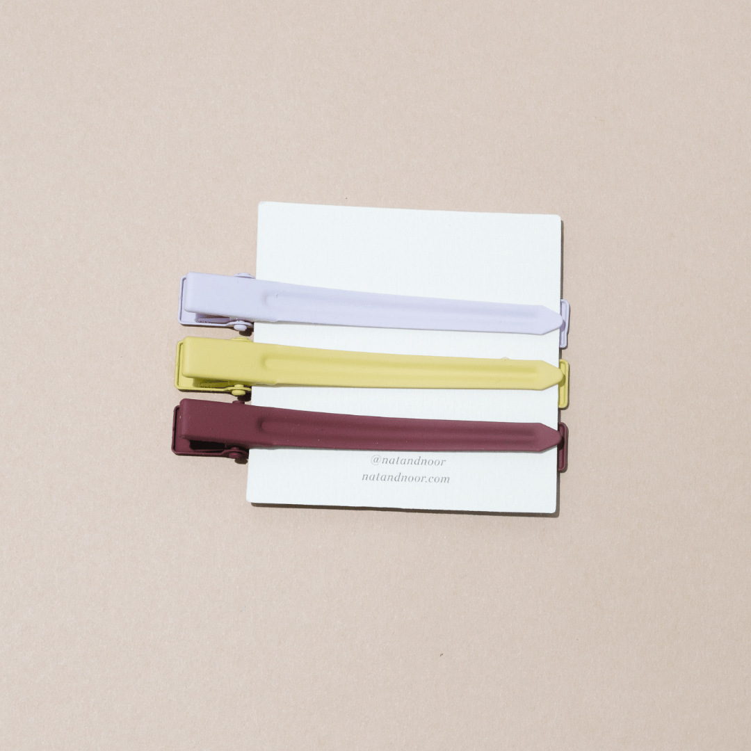 A set of three nat+noor hair clips, white yellow, and brown clipped onto a white card on a tan background