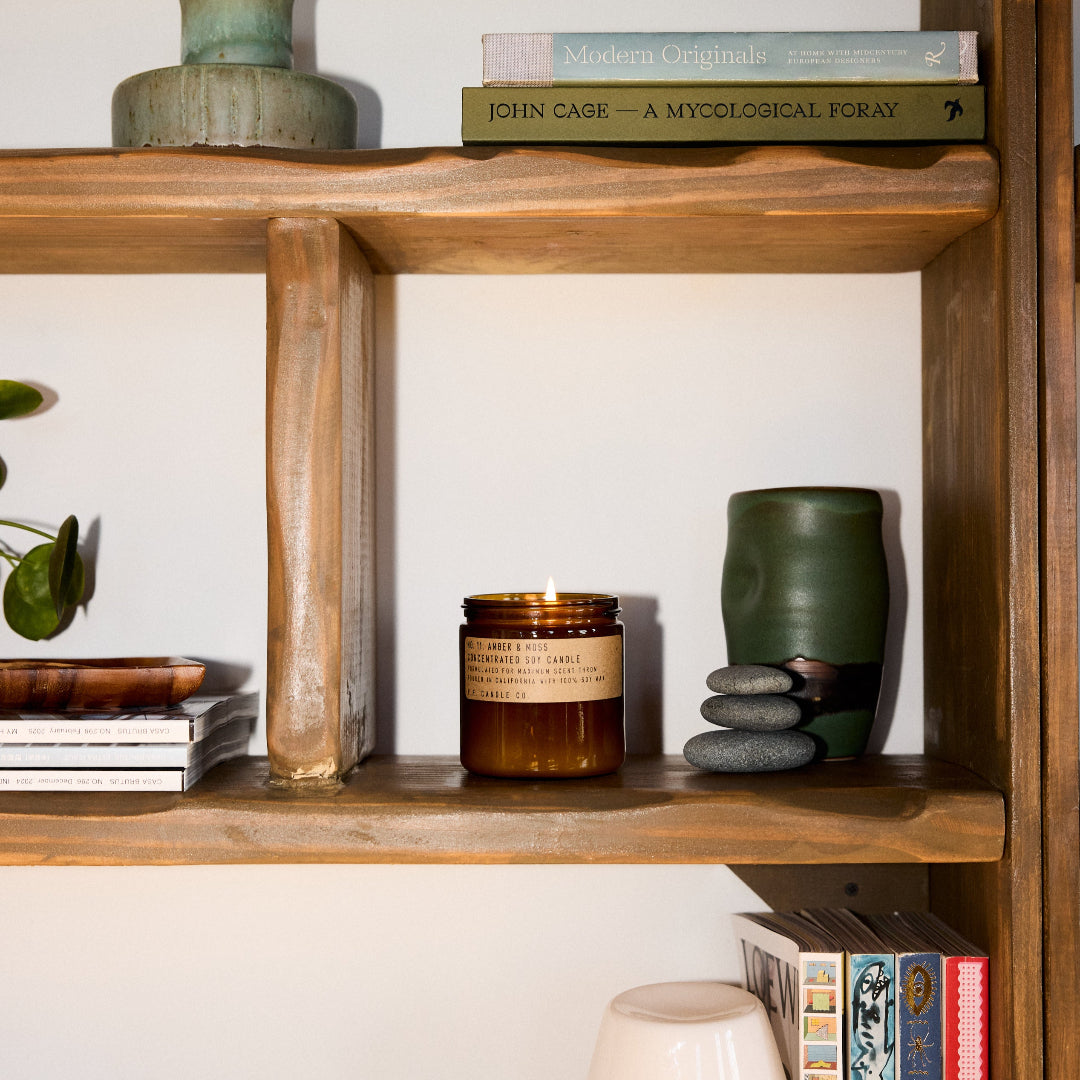 A candle with a brown label and a beige wick, placed on a wooden shelf among books, a vase, and other decorative items, with natural lighting.