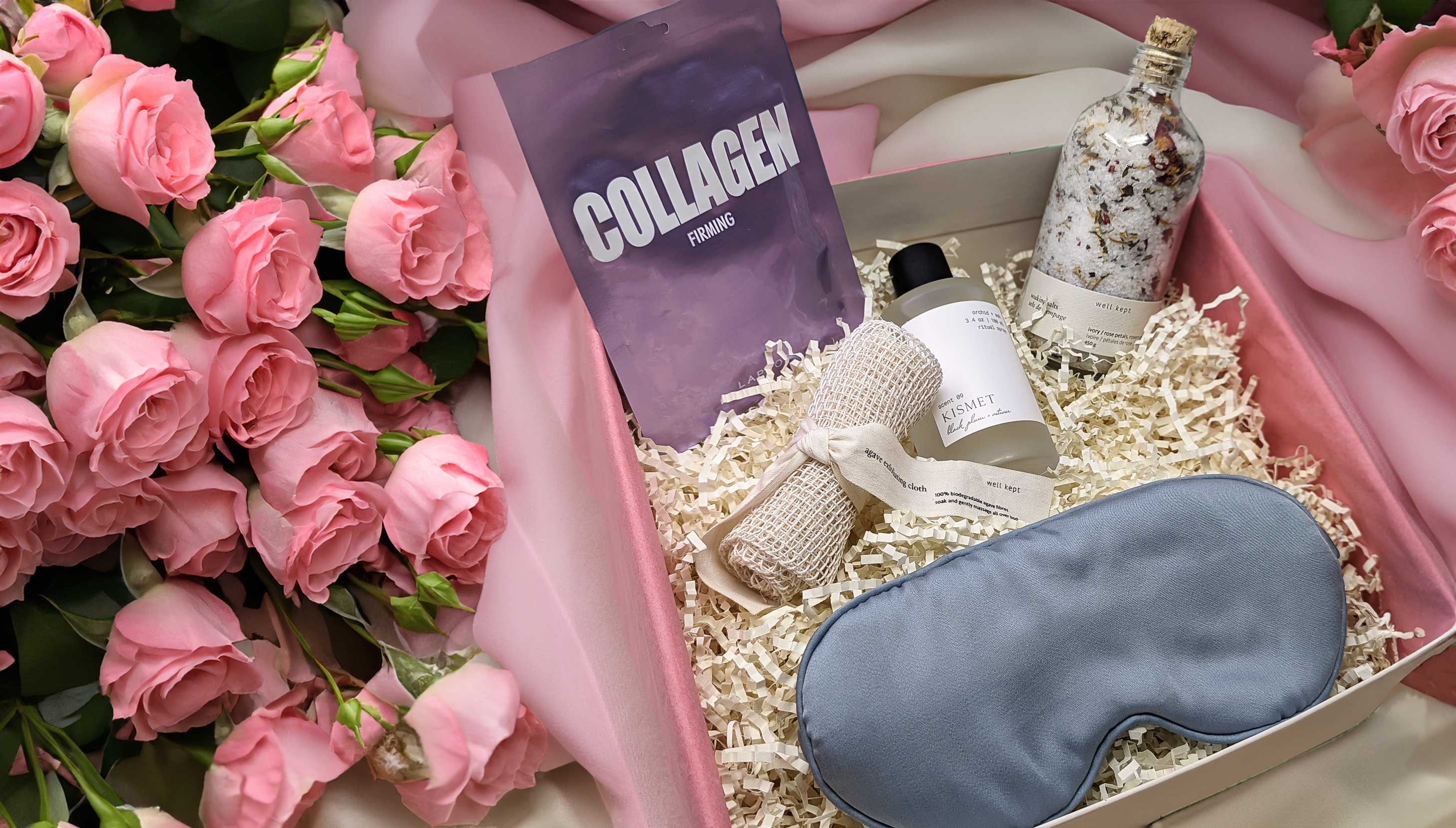 Gift box on pink cloth and pink roses. In the box is crinkle paper a sleep mask, bath salt, scrub cloth, a purple collagen firming pouch, and perfume