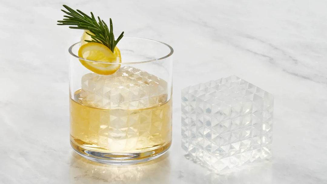 Cocktail glass half full with a W&P prism Ice cube and a twist of lemon and thyme. Drink is on a marble table next to another prism ice cube.