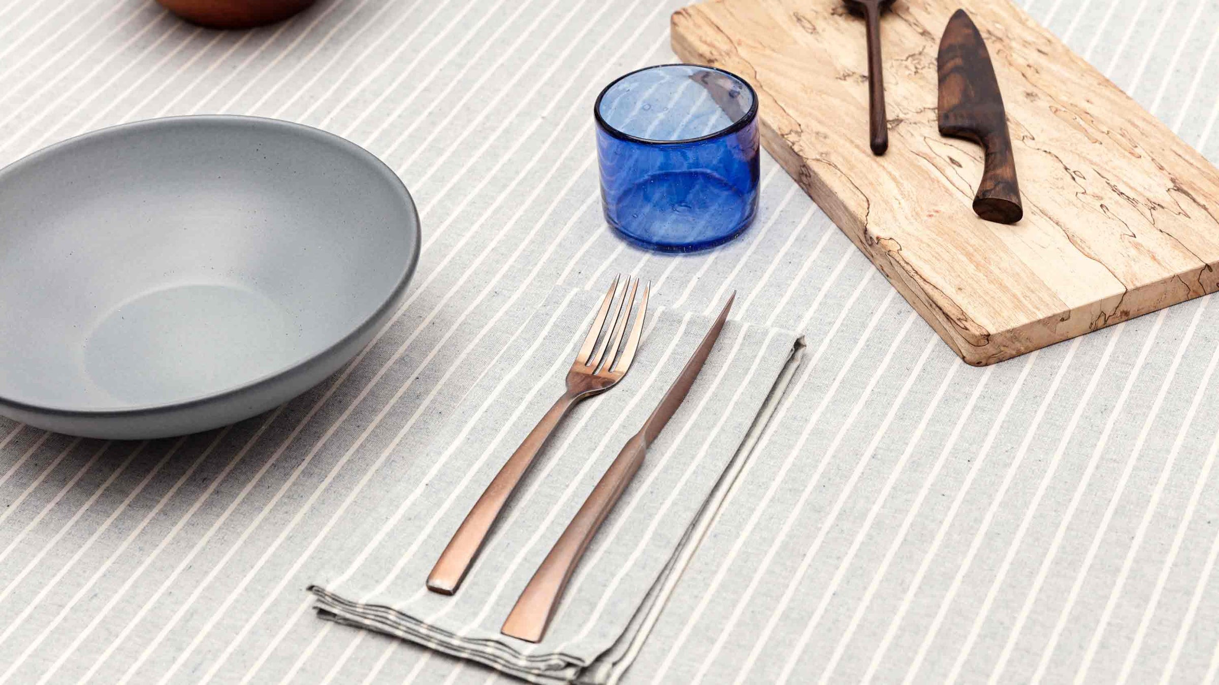 A striped tablecloth. On top is a grey ceramic bowl, a blue glass, a wooden cutting board with a dark wooden knife and spoon on top. A copper fork and knife on top of a striped napkin