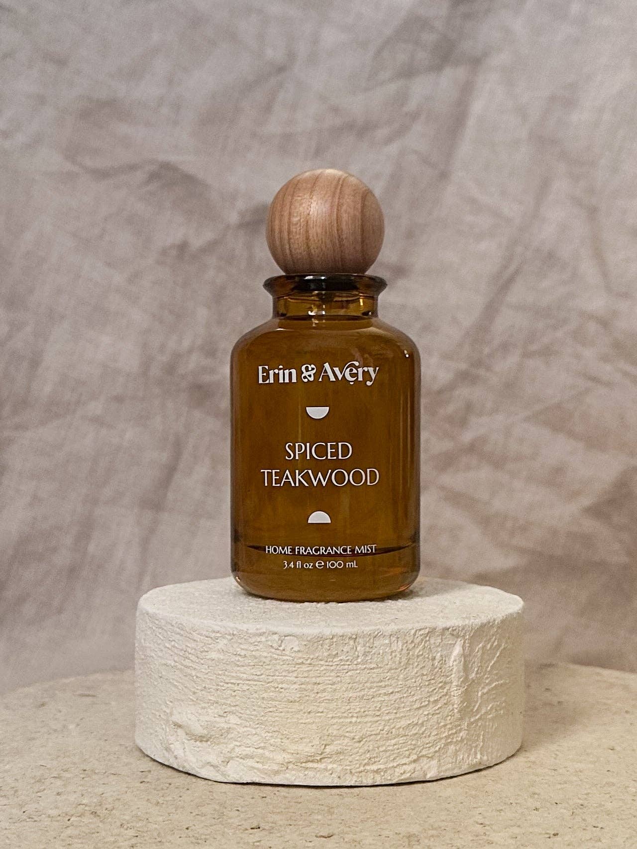 An amber glass home fragrance mist bottle with a round wooden stopper, labeled Erin & Avery Spiced Teakwood, displayed on a textured white stone pedestal against a neutral beige backdrop.
