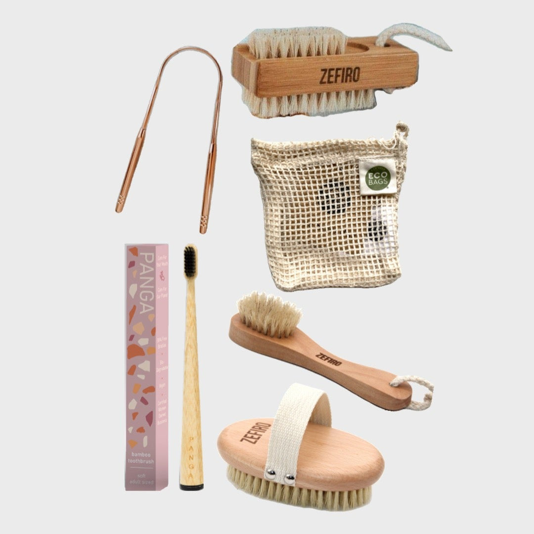 A collection of eco-friendly personal care items including a bamboo nail brush, a cotton soap bag, a rose gold stainless steel tongue scraper, a bamboo toothbrush, a face brush with natural bristles, and a dry brush with FSC-certified beechwood handle.