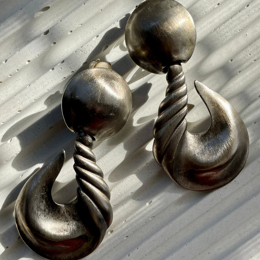 A pair of matte silver clip-on earrings with a textured design, displayed against an out-of-focus background.