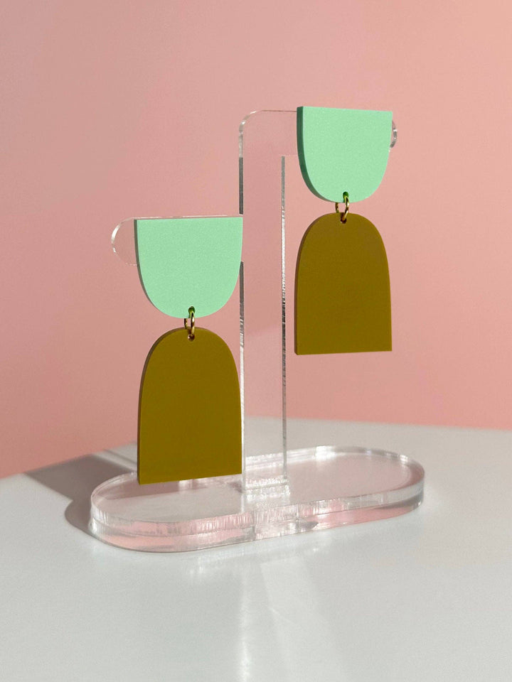 Pair of earrings with green and gold arch-shaped pendants on a clear stand against a pink background.