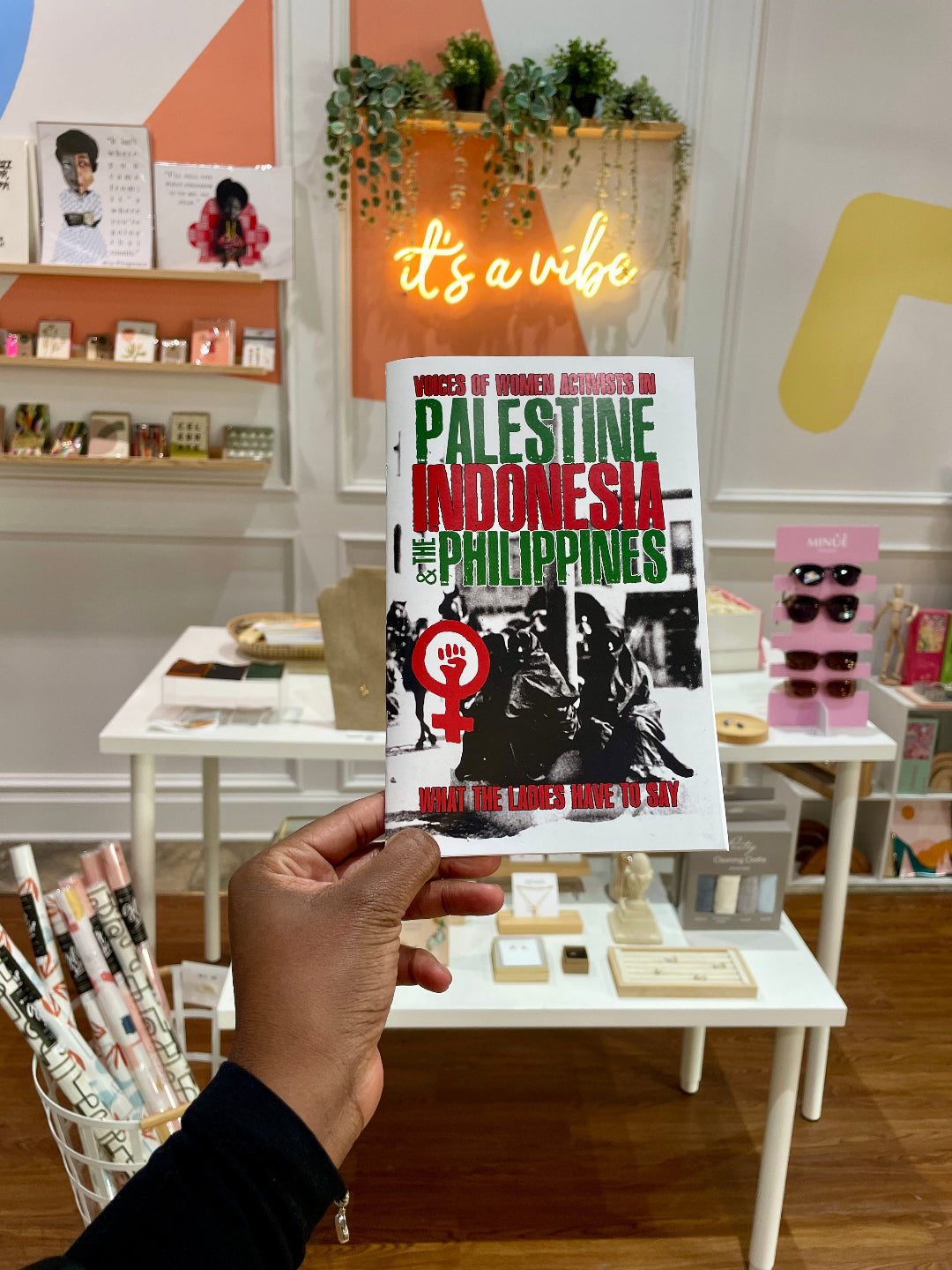 A hand holding a zine titled 'What the Ladies Have to Say: Activists in Palestine' displayed in a retail setting with various items on the shelves in the background.