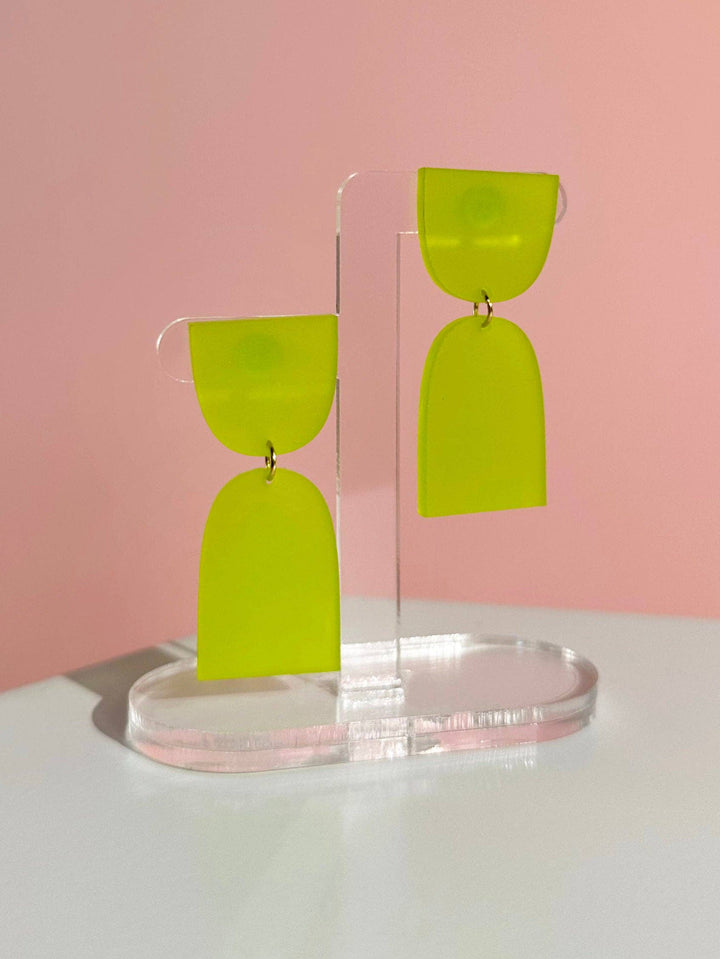 Green hourglass-shaped earrings on a clear stand against a pink background