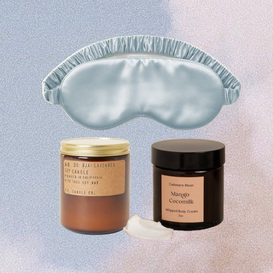 A beautifully arranged eco-friendly self-care bundle featuring a eucalyptus silk sleep mask, a soy candle in a glass jar, a travel-size mango cocomilk whipped body cream, a bamboo toothbrush, a natural bristle nail brush, and a cotton soap bag. The items are displayed on a neutral-toned background, evoking a sense of relaxation and sustainability.
