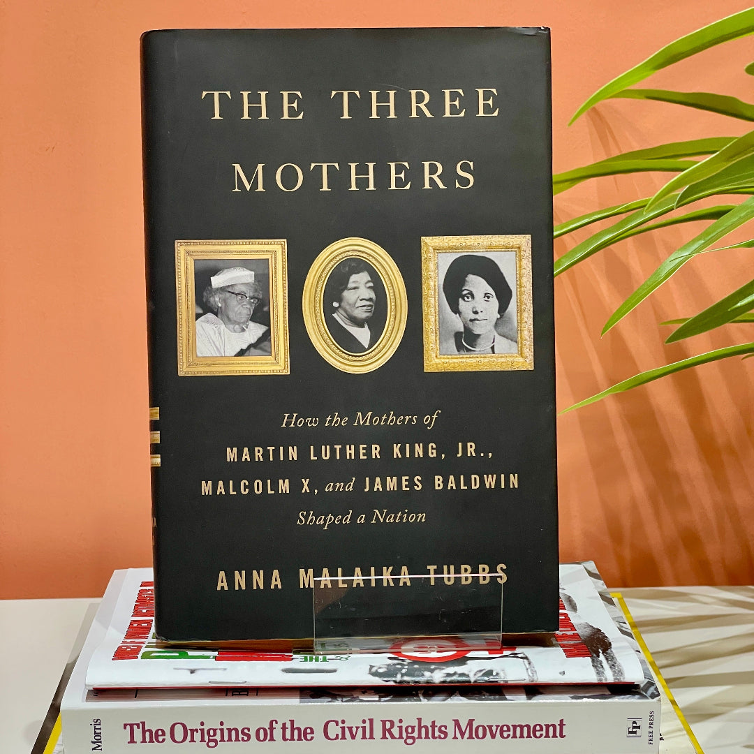 This image features a book titled "The Three Mothers: How the Mothers of Martin Luther King, Jr., Malcolm X, and James Baldwin Shaped a Nation," placed on top of a stack of other books.