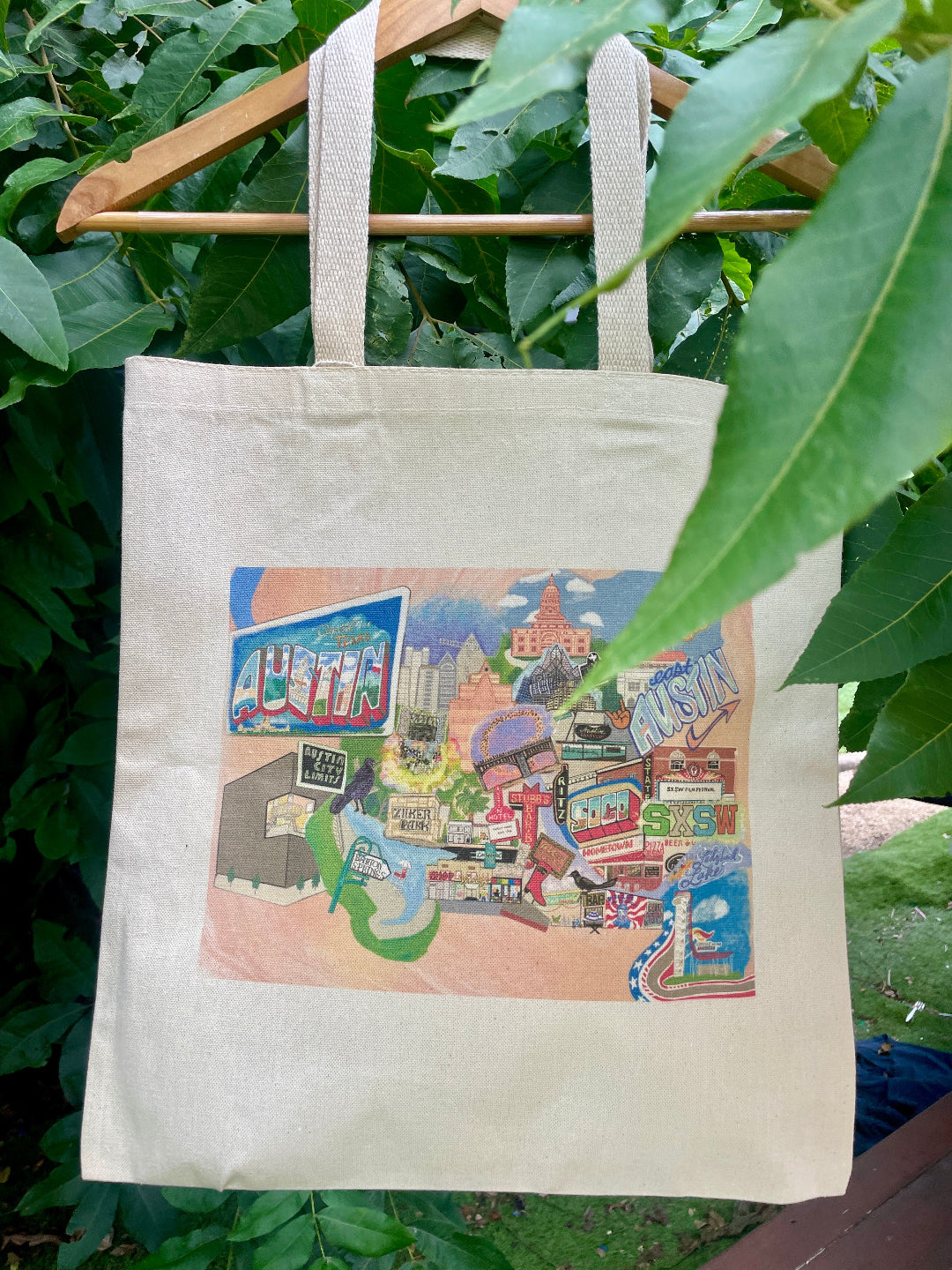 A beige cotton tote bag with hand-drawn illustrations of Austin, TX landmarks, displayed against a natural backdrop with green leaves.