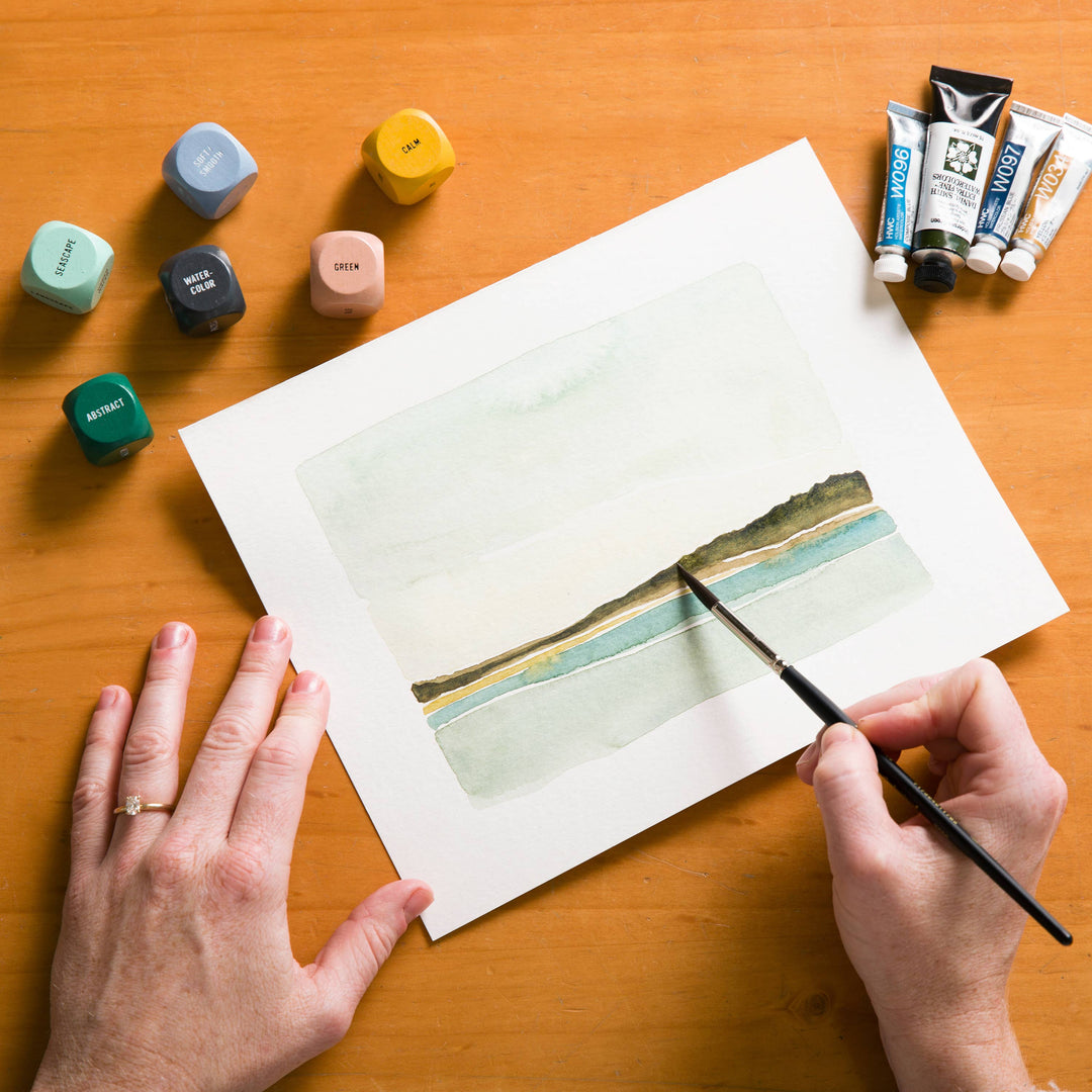 Person painting a landscape on paper with watercolor paints and brushes on a wooden table.