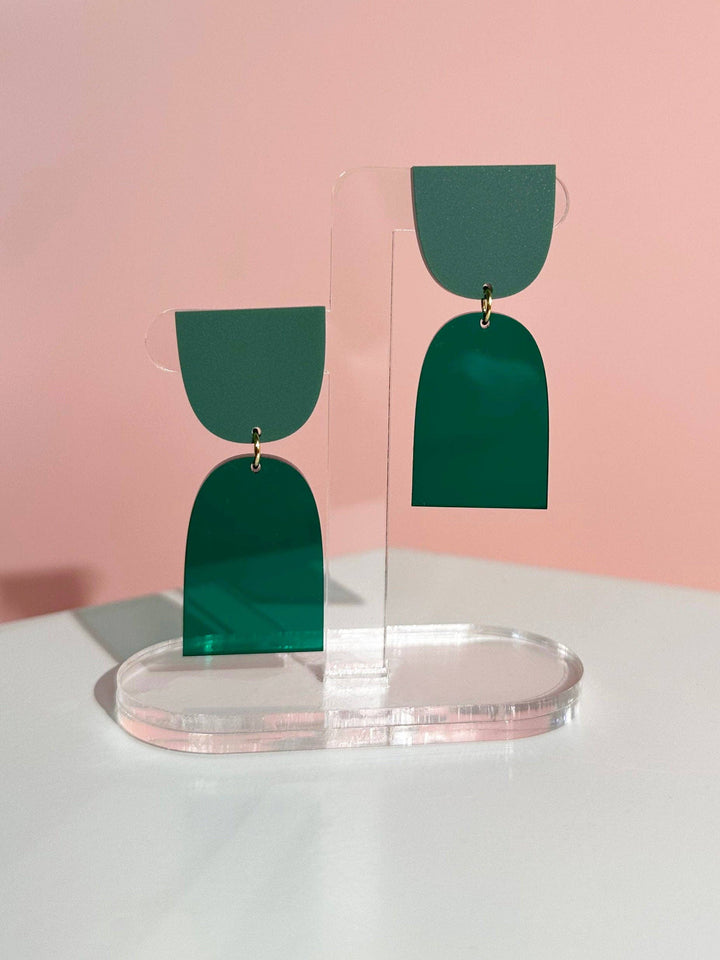 Green earrings on a clear stand against a pink background