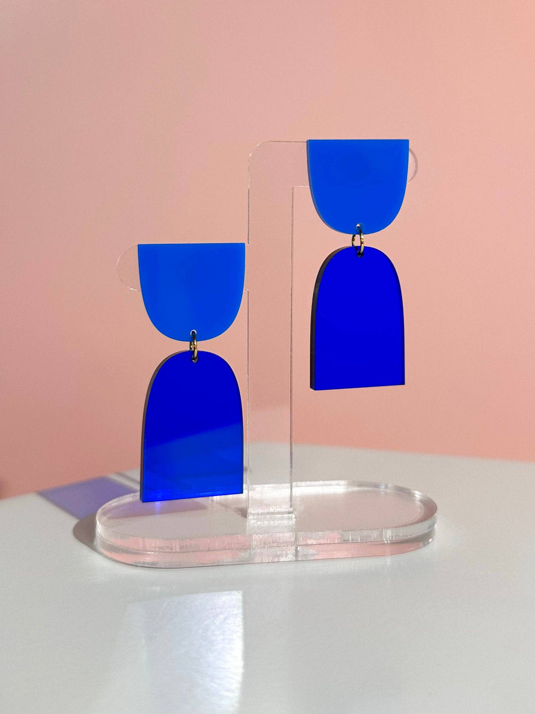 Blue earrings on a clear stand against a pink background