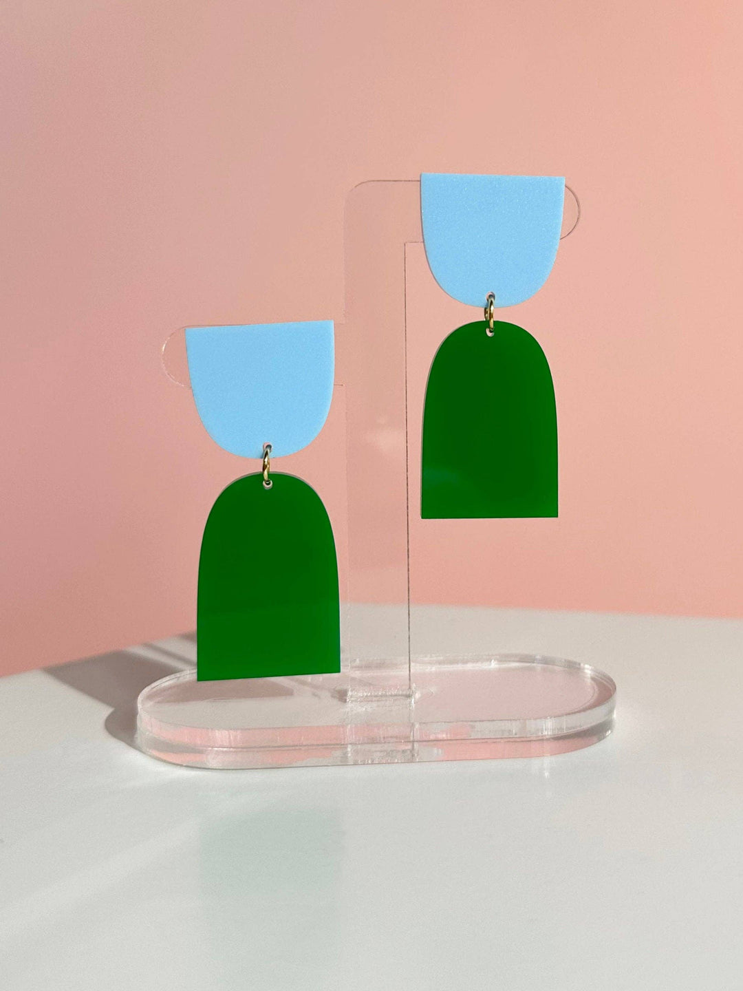 Green and blue earrings on a clear stand against a pink background