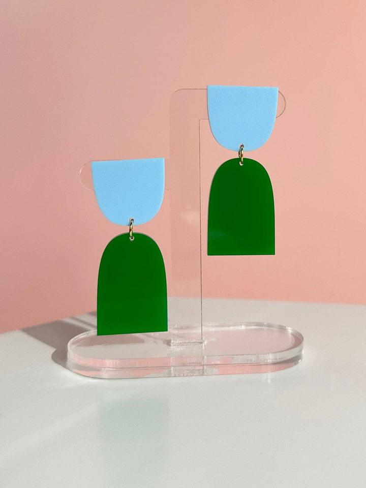 Green and blue earrings on a clear stand against a pink background