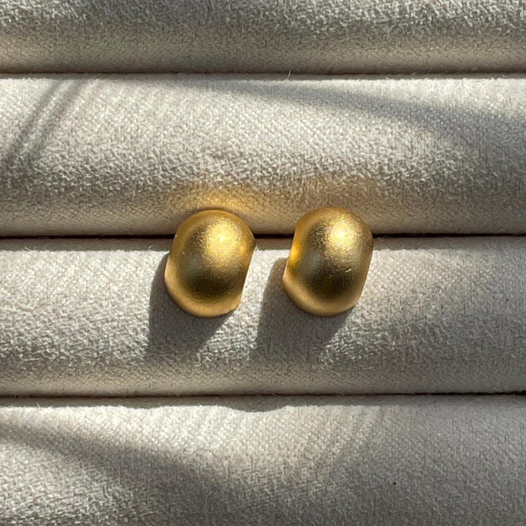 A pair of gold-colored stud earrings placed on a soft surface with a blurred background.