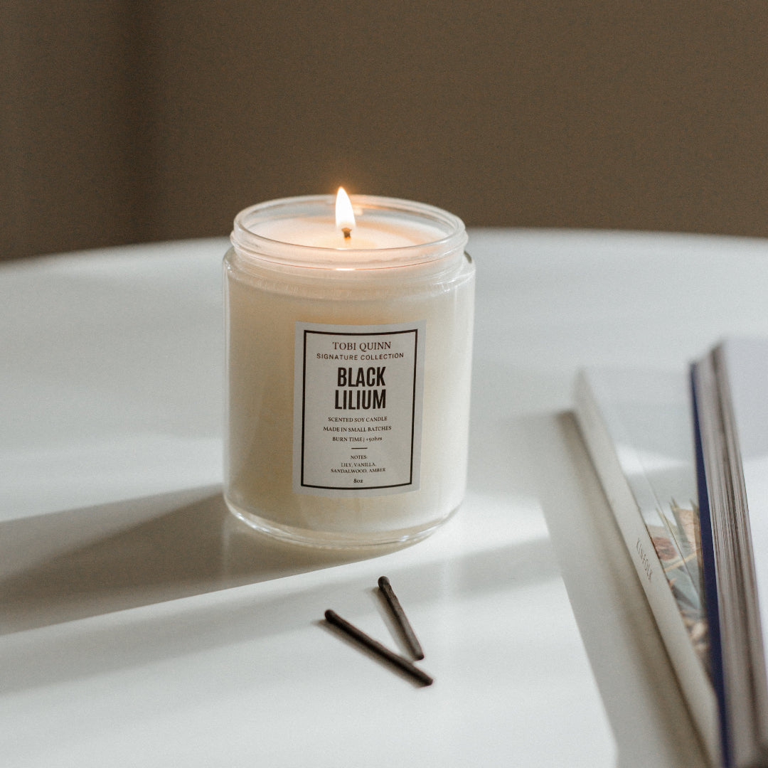 A lit soy candle labeled 'Black Lilium' in a white jar on a light background, with a wick trimming tool and printed materials nearby.