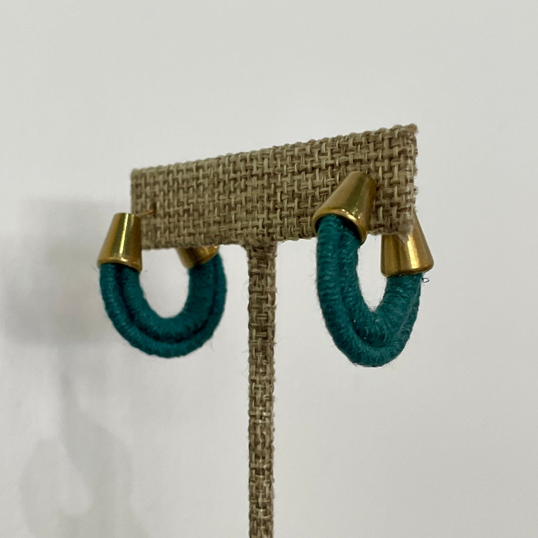 A pair of teal green hoop earrings with gold-plated hoops, wrapped in organic hemp cording, displayed on a stand.