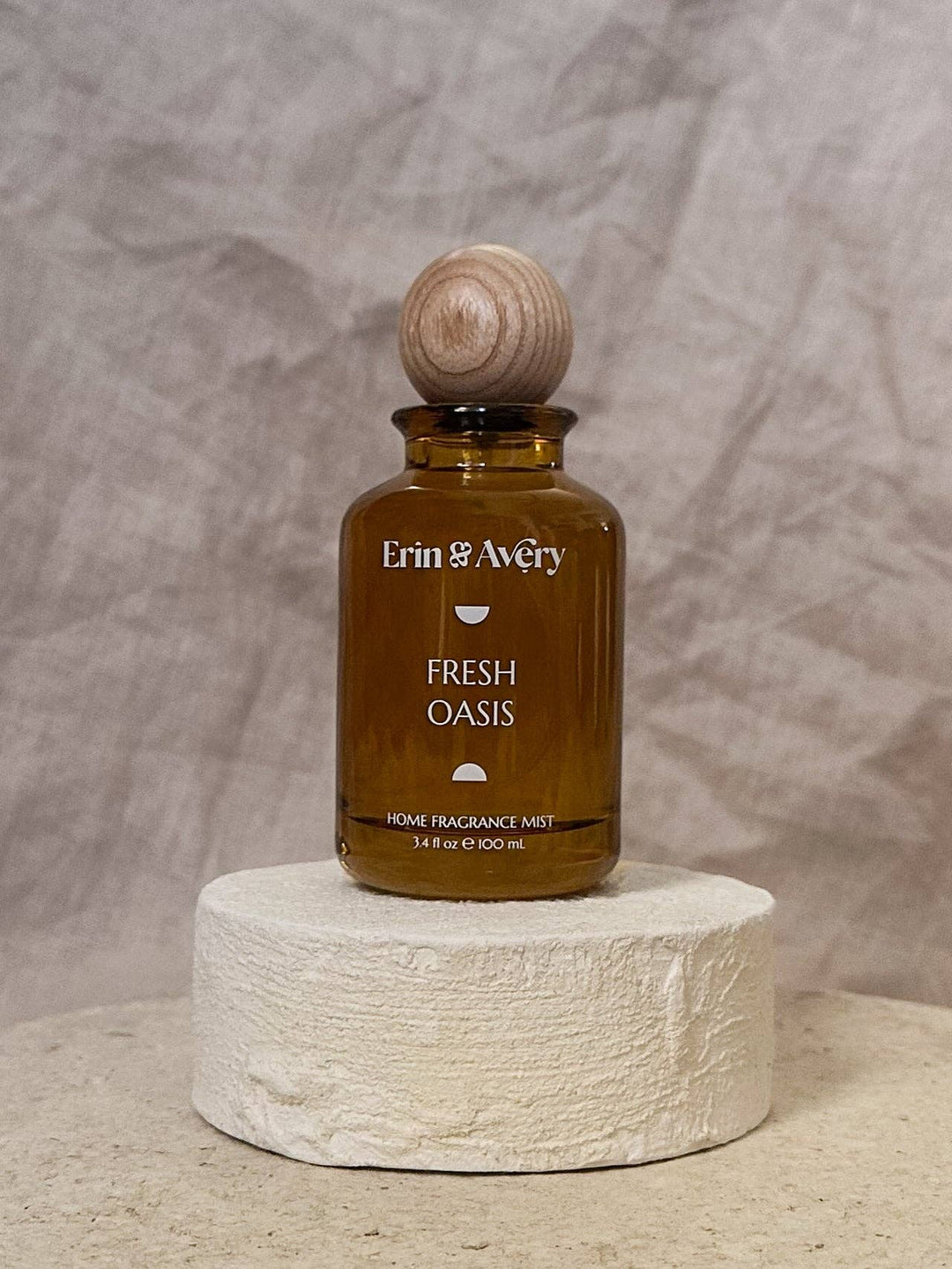 Erin &amp; Avery Fresh Oasis home fragrance mist in an amber glass bottle with a wooden stopper, displayed on a textured white stone pedestal against a neutral gray backdrop.
