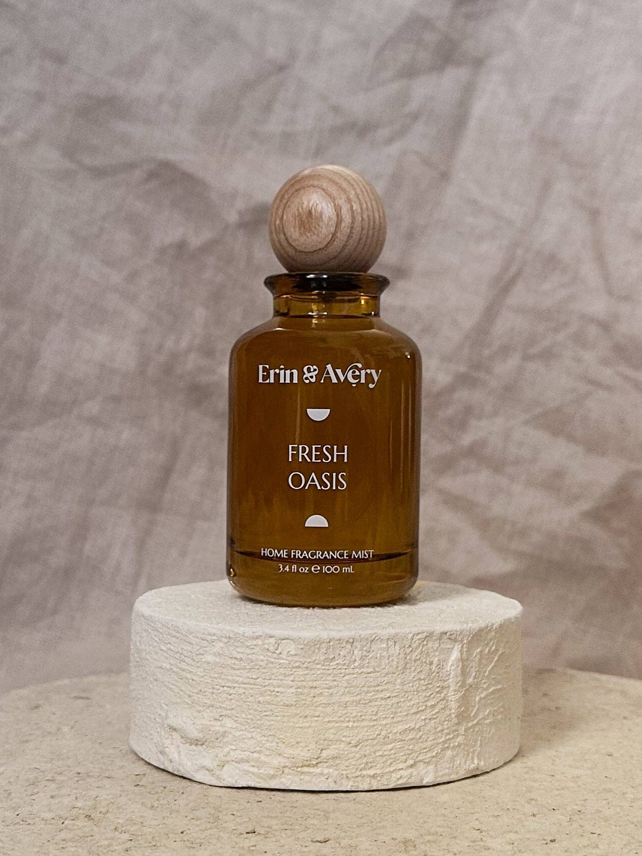 Erin & Avery Fresh Oasis home fragrance mist in an amber glass bottle with a wooden stopper, displayed on a textured white stone pedestal against a neutral gray backdrop.