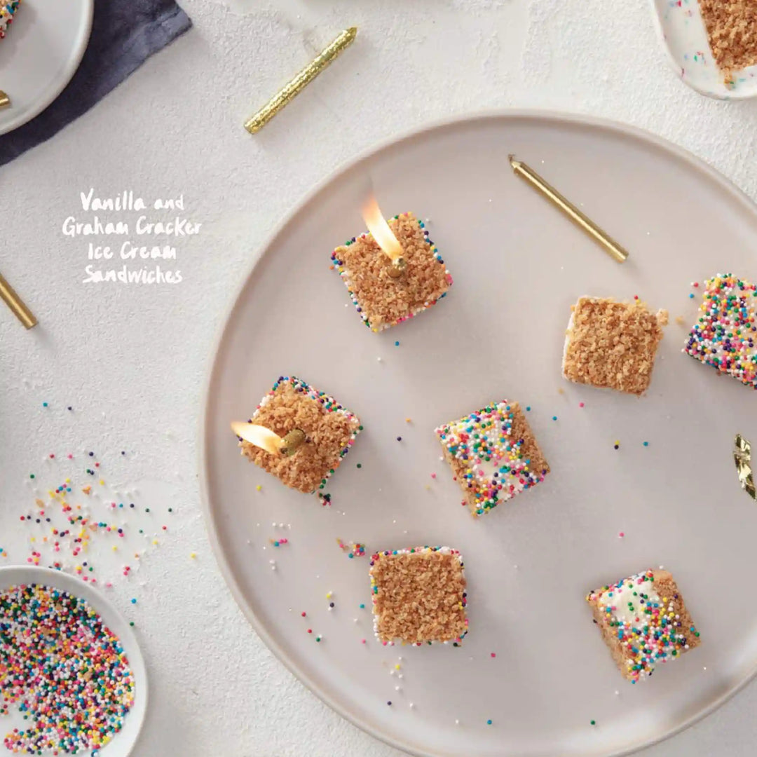 A plat of square graham cracker and ice cream treats with rainbow sprinkles. Two treats are lit with birthday candles.