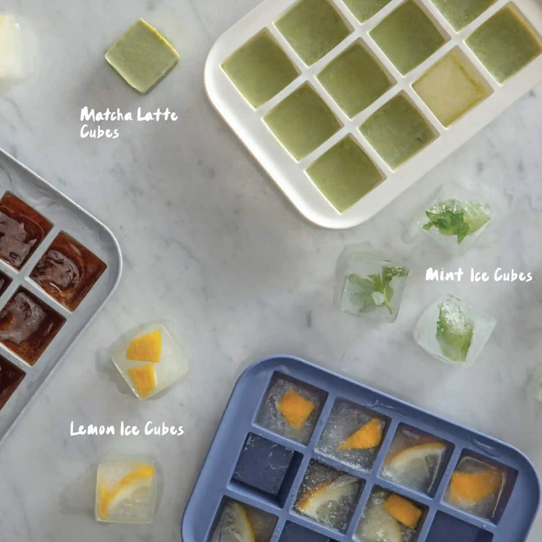 A grey, a white, and a blue ice cube tray holder on a marble background. There are ice cubes on the marble as well. Each ice cube tray has different ice cubes in it. 