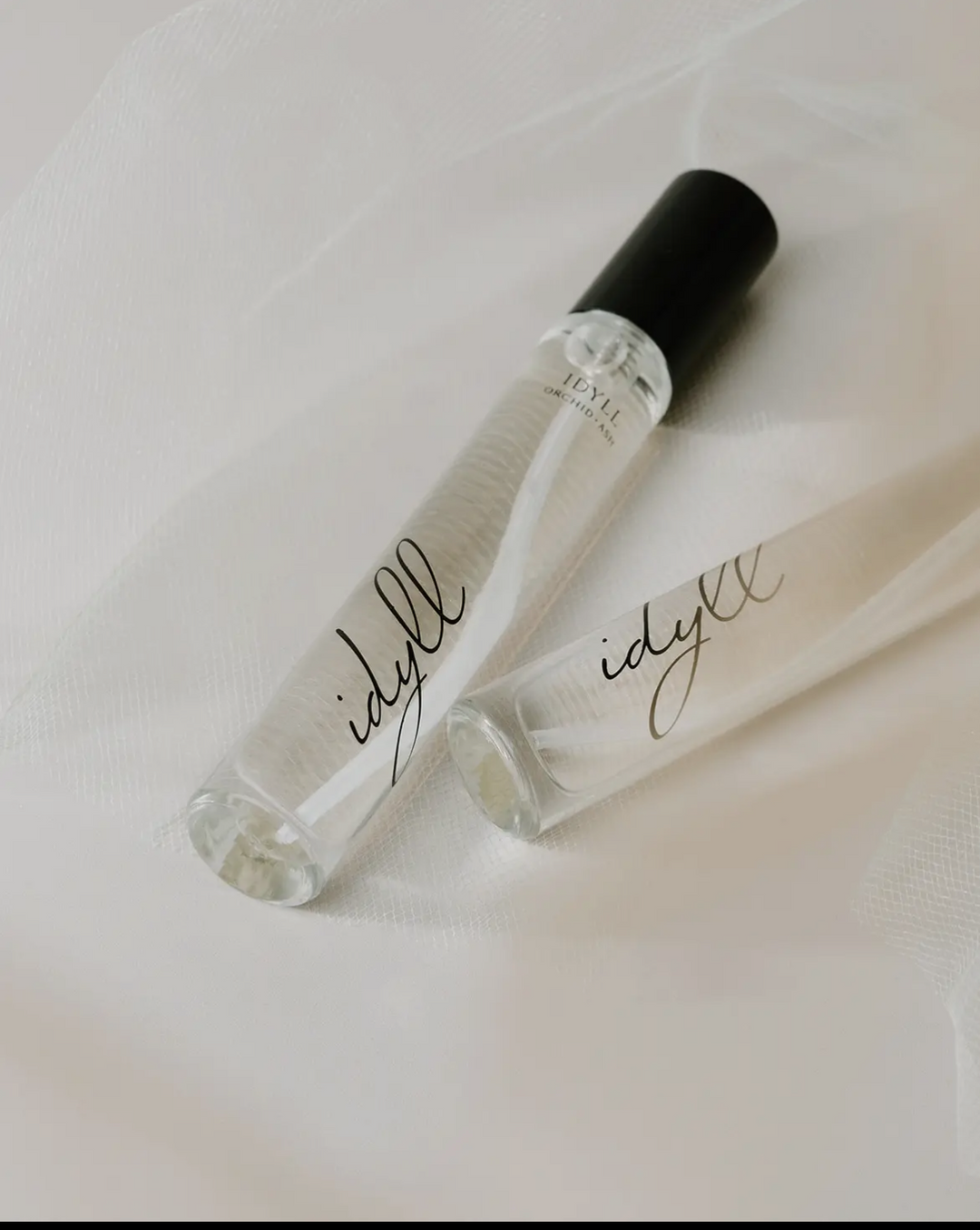 Two clear 'idyll' perfume bottles with black caps on a white fabric background