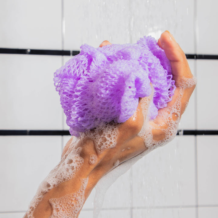 Hand holding a purple bath sponge with soap suds against a tiled bathroom wall.