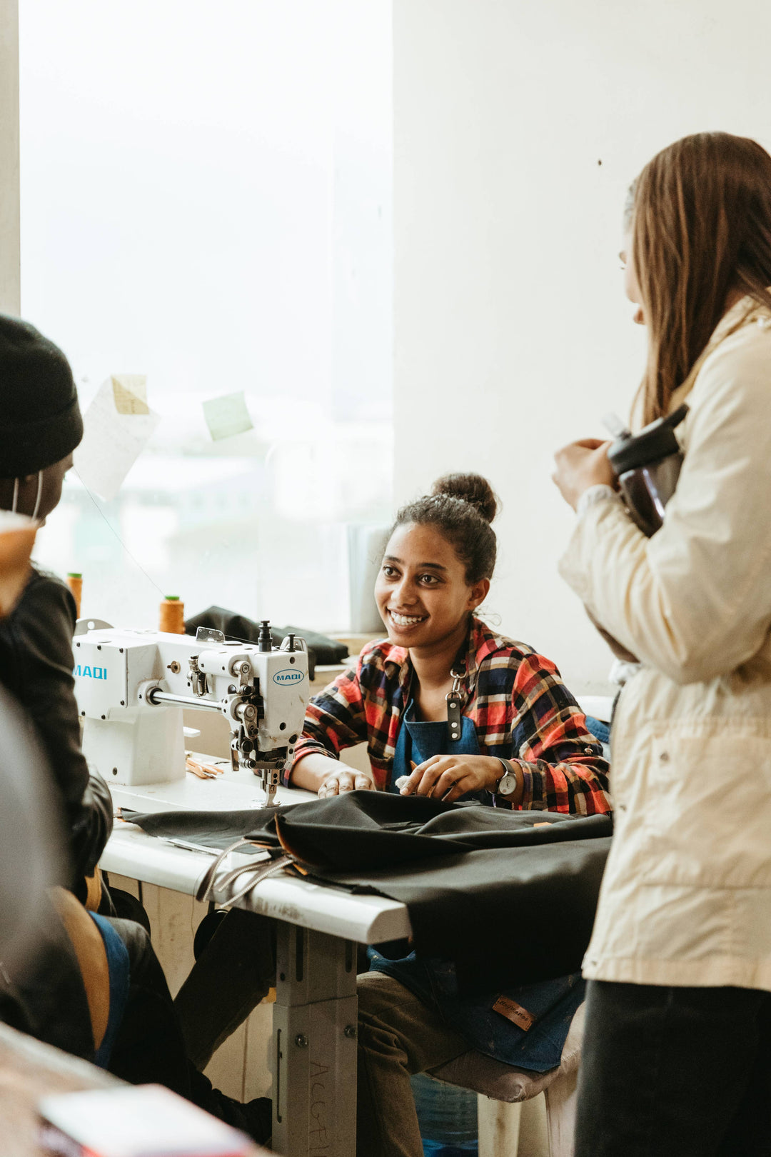 Person sitting at a sewing machine with another person standing nearby in a workshop setting.