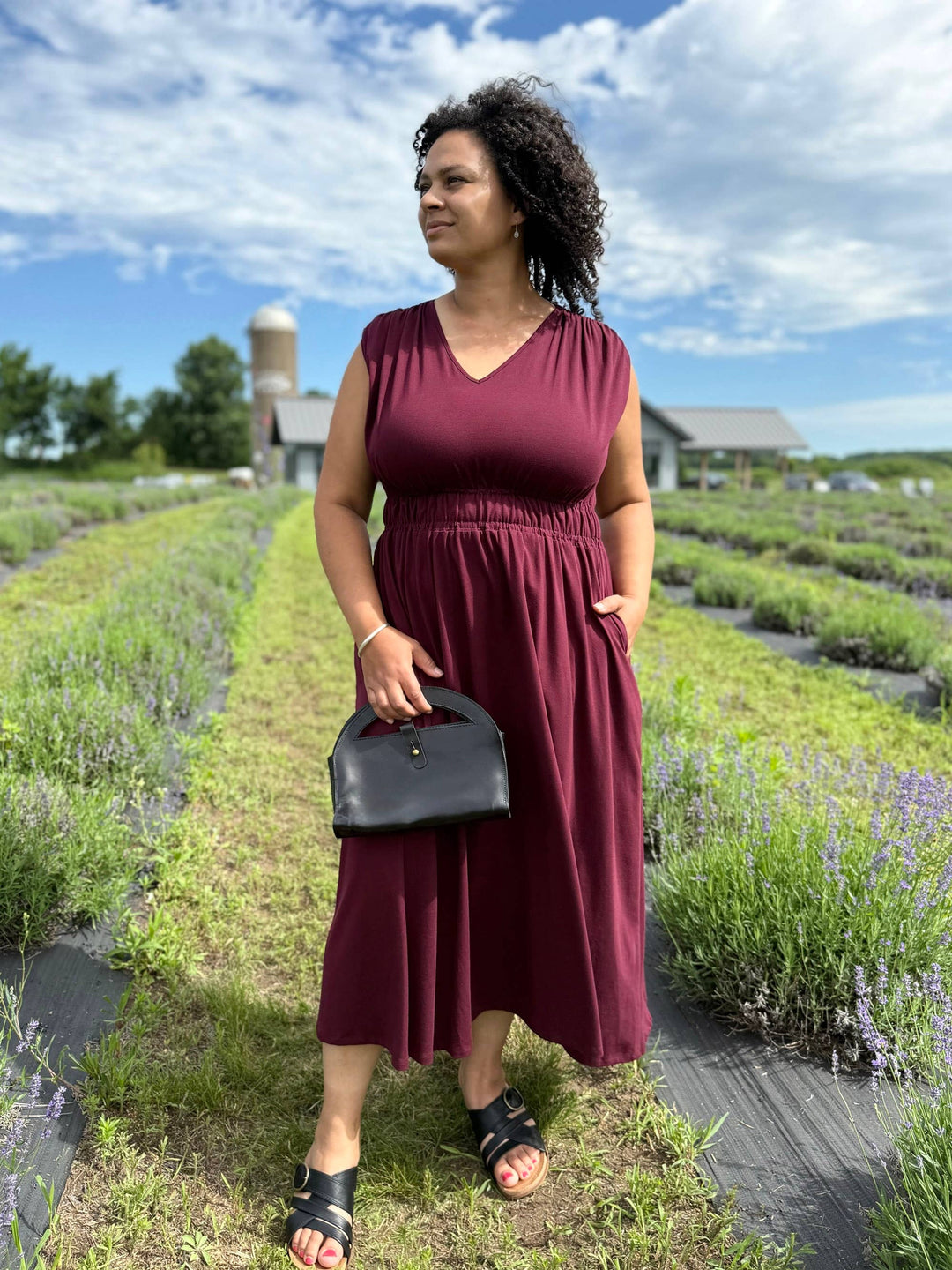 Woman in a burgundy dress standing in a lavender field with a black handbag.