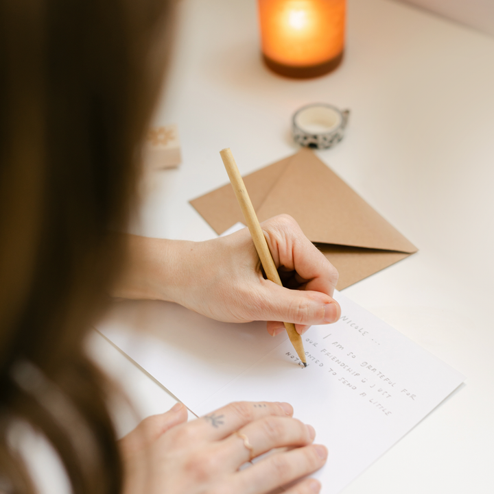 Person writing on a piece of paper with a pencil, surrounded by a candle and envelope on a table.