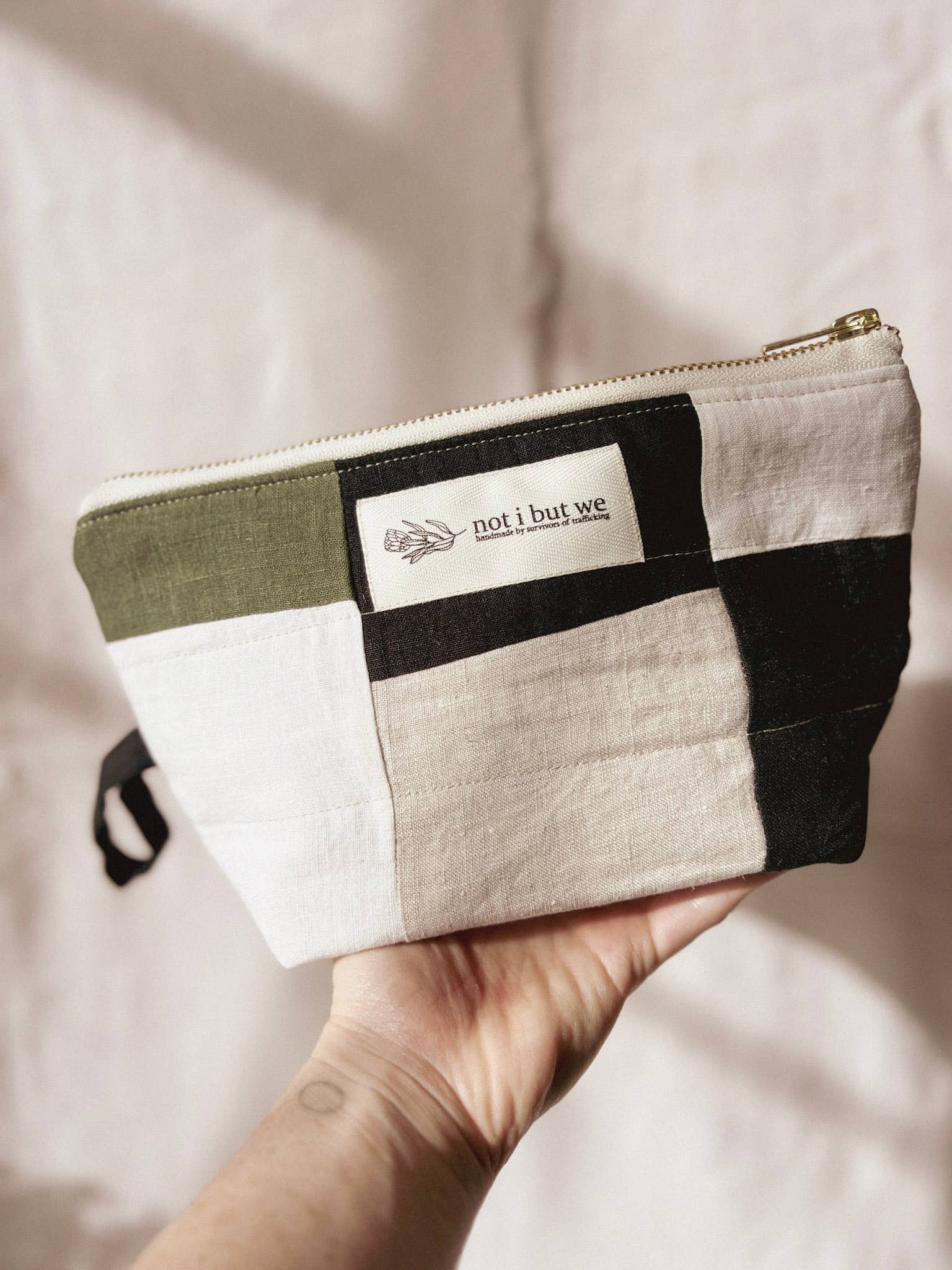 Hand holding a patchwork pouch with 'not i but we' branding against a neutral background