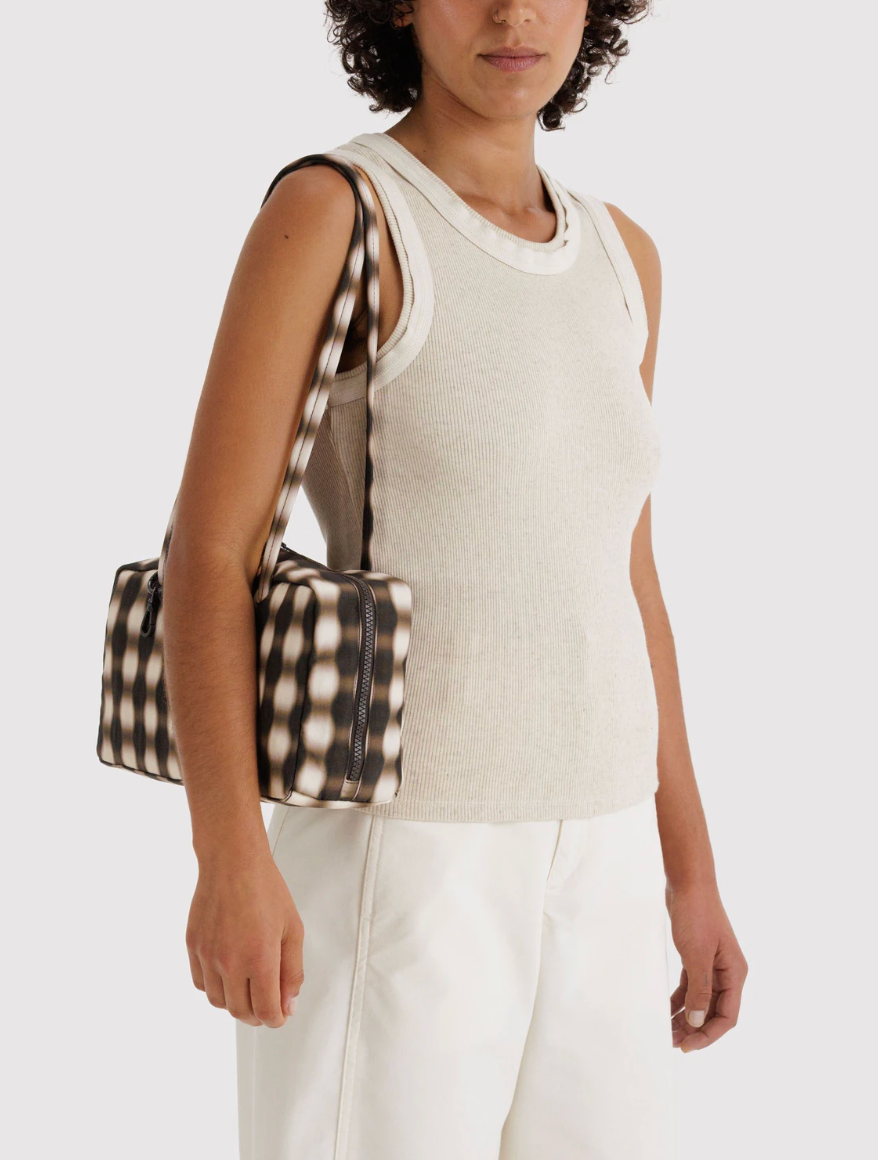 person in light top and bottom holding Nylon Bowler Bag Blurred Gingham Brown on right shoulder against white backdrop