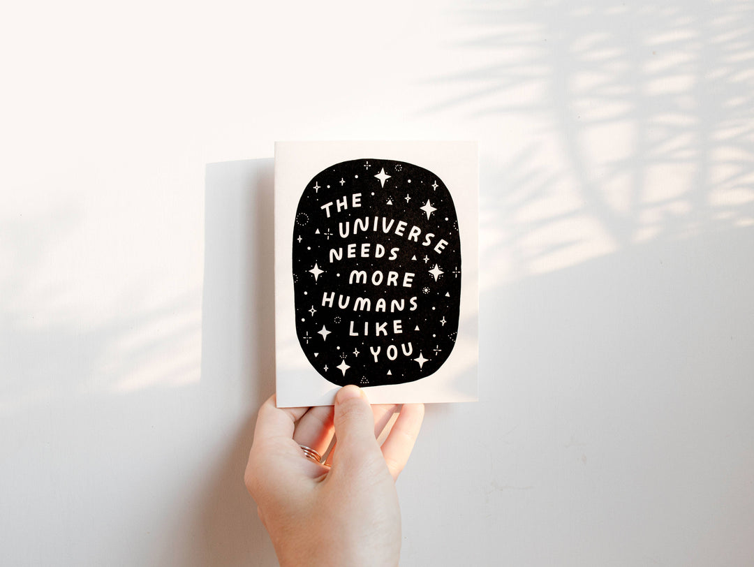 Hand holding a card with 'The universe needs more humans like you' text against a light background