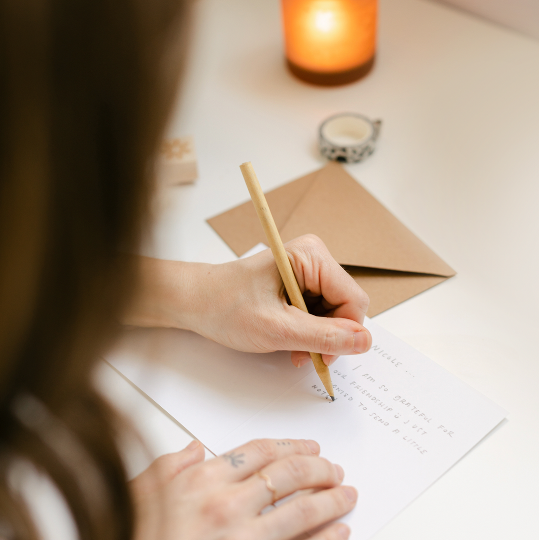 Person writing on a piece of paper with a pencil, surrounded by a candle and envelope on a table.